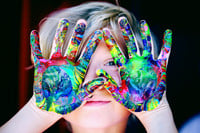 Elementary student with multiple colors of paint on their hands, holding them in front of their face